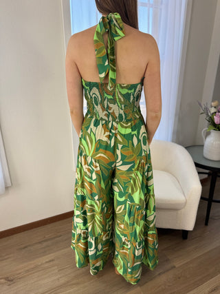Person wearing a green patterned dress standing in a room with a white chair and a small table with a plant.