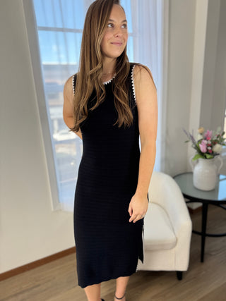 Woman in a black dress standing in a room with a window and a small table with flowers.