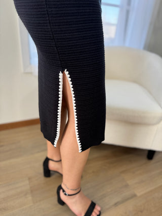 Person wearing a black ribbed dress with a high slit in a room with a white chair and wooden floor.