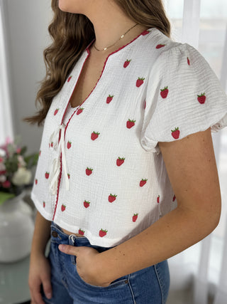 Woman wearing a white blouse with red strawberry pattern and blue jeans indoors.