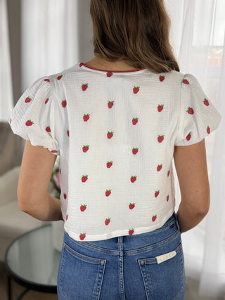 Person wearing a white blouse with red strawberry pattern and blue jeans indoors.