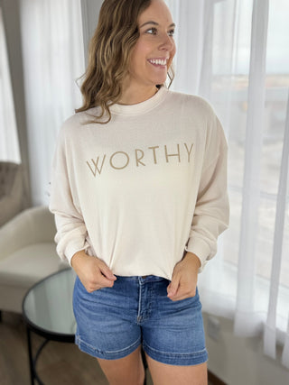 Woman wearing a beige sweatshirt with 'WORTHY' text and denim shorts indoors.