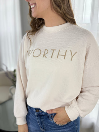 Person wearing a beige sweater with 'WORTHY' text, standing indoors.