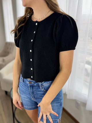 Person wearing a black short-sleeve button-up top and blue jeans indoors.