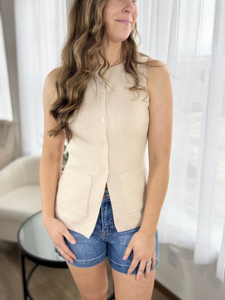 Woman wearing a beige vest over a white shirt with denim shorts indoors.