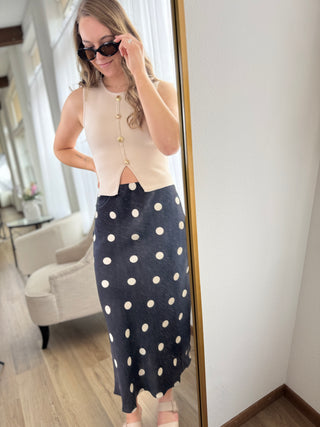Woman wearing a beige cardigan and navy polka dot skirt in front of a mirror.