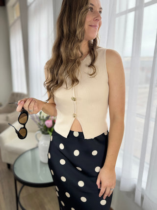 Woman holding sunglasses indoors, wearing a cream sleeveless top and black polka dot skirt.
