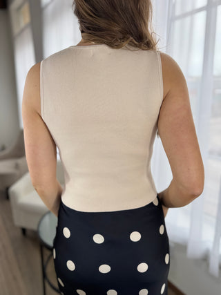 Person wearing a beige sleeveless top and black polka dot skirt indoors.