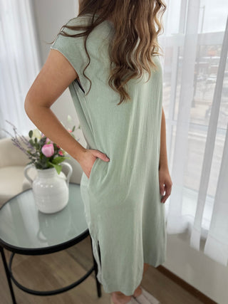 Woman wearing a light green dress standing in a bright room with white curtains.