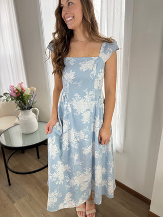 Woman wearing a blue floral dress in a room with a table and flowers.