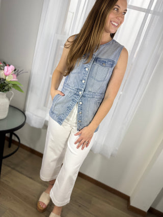 Woman wearing a denim sleeveless shirt and white pants indoors.