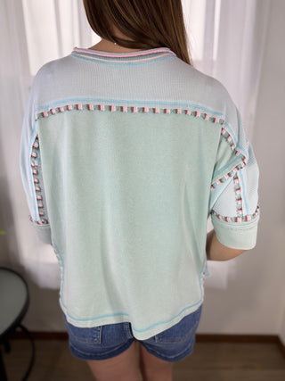 Light blue sweater with patterned trim worn by a person against a white curtain background