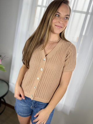 Woman wearing a beige cardigan and denim shorts indoors with white curtains in the background