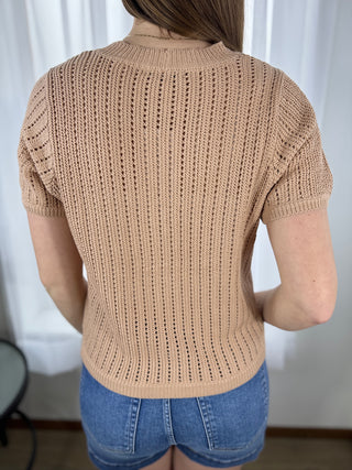 Person wearing a beige knitted short-sleeve sweater and blue shorts against a white curtain background