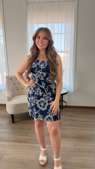 A woman modeling a navy blue strapless mini dress with a white floral pattern, standing in a brightly lit room with wood floors and a window in the background.