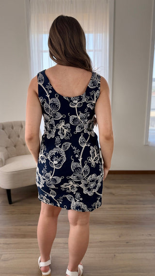 A woman viewed from behind wearing a sleeveless navy blue dress with a white floral pattern, standing in a brightly lit room with light wood floors and a cream tufted chair in the background.