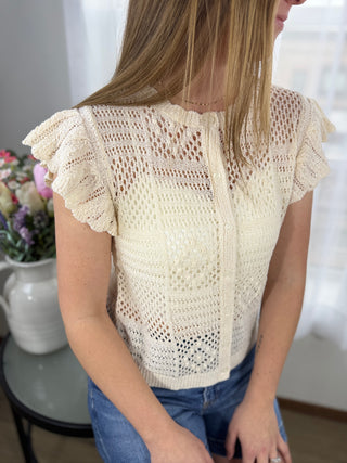 Woman wearing a beige crochet top sitting indoors with a blurred background