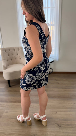 A woman wearing a sleeveless navy blue and white floral print mini dress, shown from the back in a brightly lit room with wood floors and a tufted armchair.