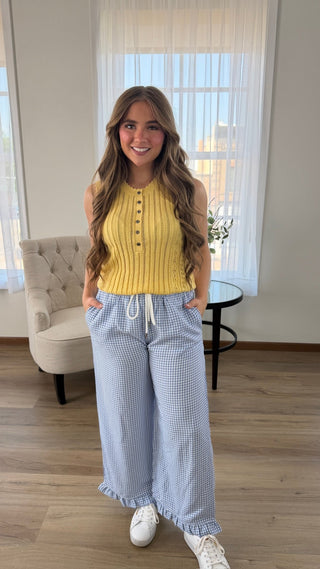 A woman standing in a bright room wearing a sleeveless yellow ribbed knit top with buttons and light blue-and-white gingham wide-leg pants, paired with white sneakers.