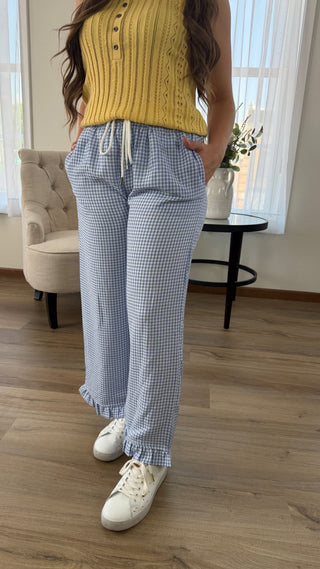 A person wearing loose blue-and-white gingham drawstring pants with a yellow ribbed knit top and white sneakers, standing in a bright indoor room with wood flooring, a chair, and a small table.
