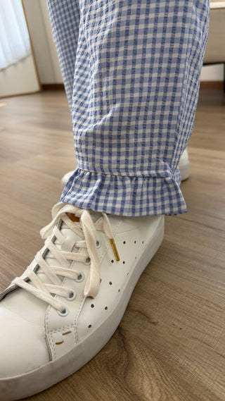 A close-up of a person's foot wearing a white leather low-top sneaker with white laces, paired with blue and white gingham checkered pants, standing on a light wood floor.