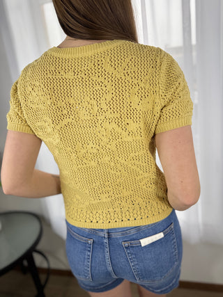Person wearing a yellow knitted top and blue shorts against a white curtain background