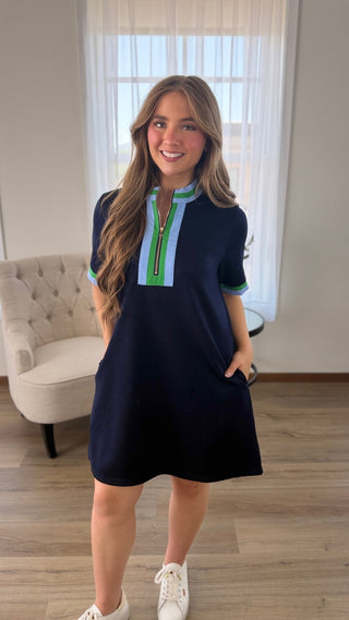 A woman modeling a short-sleeve navy blue dress with a light blue and green zipper-style placket, standing in a bright room with wood floors and a white tufted armchair.
