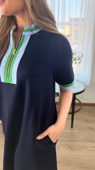 A woman wearing a short-sleeve navy dress with a light blue and green striped collar and sleeve trim, standing in a bright indoor room.