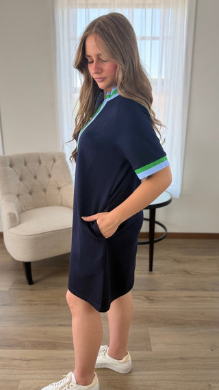 A woman modeling a navy blue short-sleeve dress with light blue and green striped accents on the collar and sleeve cuffs, standing in a bright room with wood floors and a tufted armchair.