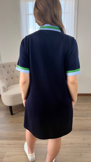 A woman viewed from behind wearing a short-sleeve navy polo dress with green and light blue striped accents on the collar and sleeve cuffs, standing in a room with light wood floors and a beige armchair.