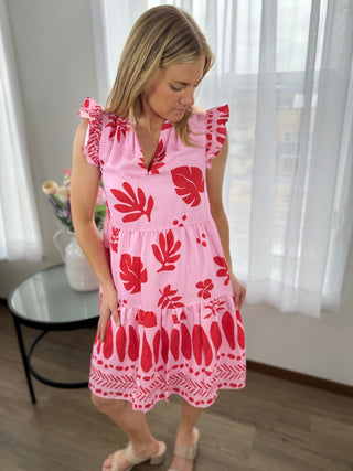 Woman wearing a pink dress with red floral patterns in a room with white curtains.