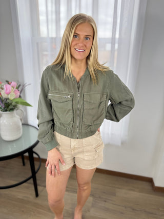 Woman wearing a green jacket and beige shorts indoors with a blurred background