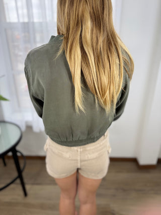 Person wearing a green jacket and beige shorts in a room with white curtains.