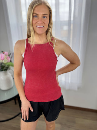 Woman wearing a red tank top and black shorts indoors with white curtains in the background