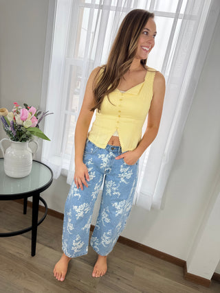 Woman wearing a yellow sleeveless top and blue floral pants standing in a room with a table and flowers.