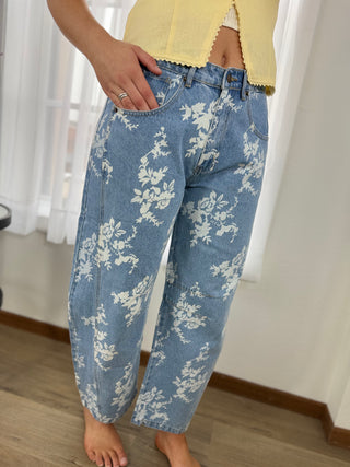 Person wearing blue floral-patterned jeans indoors with white curtains in the background