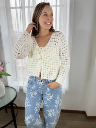Woman wearing a white crochet cardigan and blue floral pants indoors.