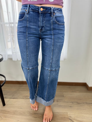 Person wearing blue jeans standing on a wooden floor with white curtains in the background