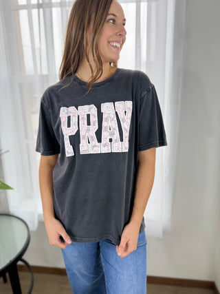 Woman wearing a black t-shirt with 'PRAY' printed on it, standing indoors.