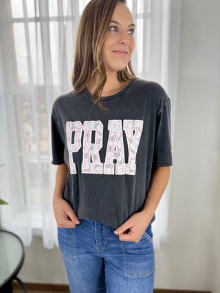 Woman wearing a dark gray t-shirt with 'PRAY' printed on it, standing indoors.