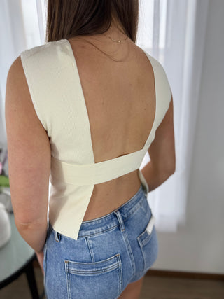 Person wearing a cream sleeveless top with cut-out design and blue jeans indoors.