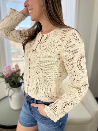 Woman wearing a beige crochet sweater and blue jeans indoors.