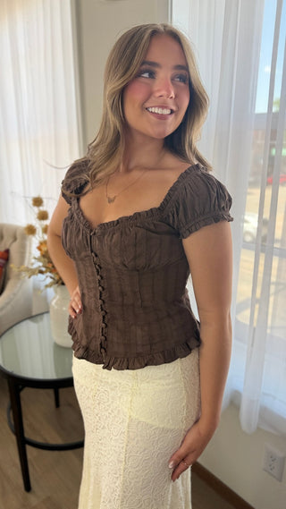 A woman wearing a brown ruffled off-the-shoulder top and a cream lace skirt, standing in a brightly lit room with white curtains.
