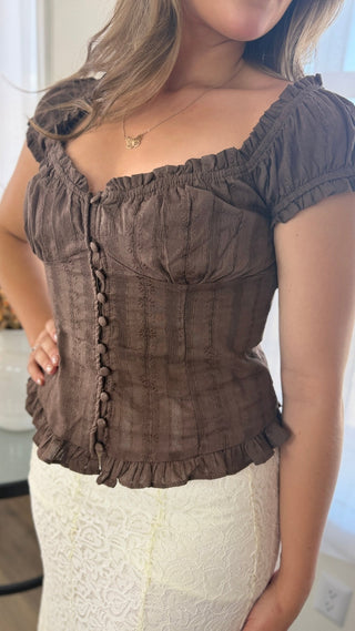 A woman wearing a brown short-sleeve ruffled top with a buttoned front and a white lace skirt, photographed indoors.