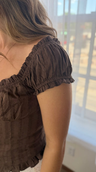 A close-up lifestyle shot of a woman wearing a dark brown short-sleeve top with ruffled trim, standing near a window.