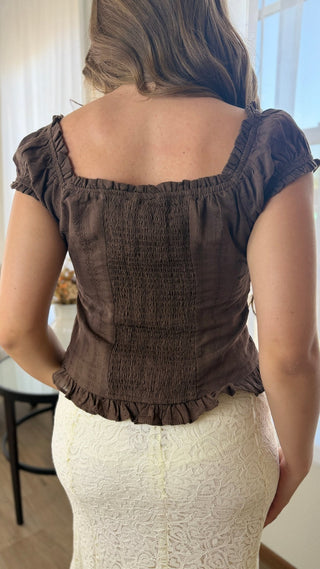 A woman shown from the back wearing a dark brown smocked short-sleeve top with a square neckline and ruffled trim, paired with a cream lace skirt.