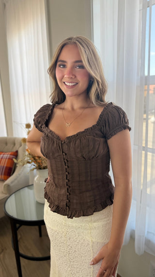 A woman wearing a brown ruffled short-sleeve top and a cream lace skirt, standing in a bright room with sheer curtains.