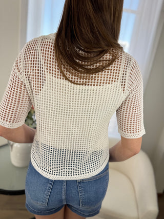 Person wearing a white mesh top and denim shorts in a room with light-colored walls.