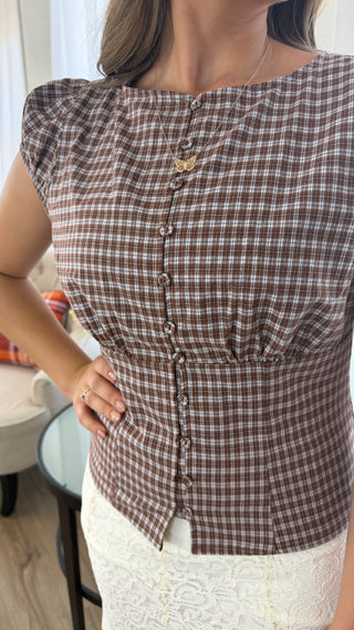 A woman wearing a brown and beige gingham pleated top with a button-down front, paired with a white lace skirt, in an indoor setting.