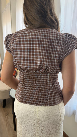 A woman viewed from behind wearing a brown and beige gingham smocked blouse with puffed cap sleeves, paired with a cream lace skirt.